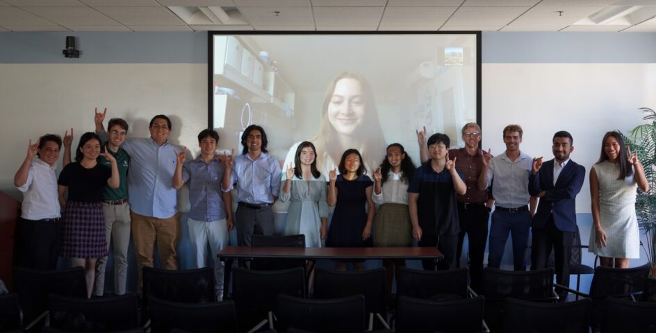 Fourteen students standing in front of a screen, showing the fifteenth student on zoom, with hands raised for the UCI zot.