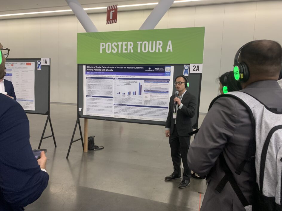 Chandler stands with a microphone, talking to people in front of a large poster, titled "Effects of Social Determinants of Health on Health Outcomes Among Patients with Obesity.