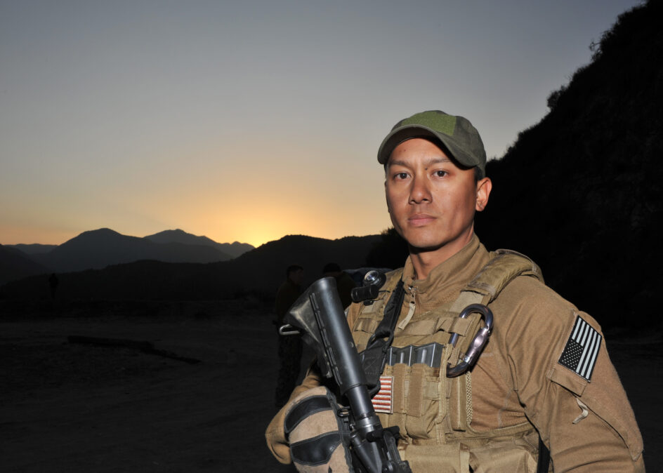 Balacy in combat gear holding gun, with hills in the backdrop.