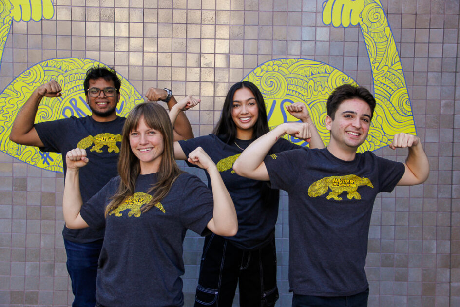 Students standing in front of a wall at UCI with artwork portraying two strong arms. The students are raising their arms as well, showing their strength.