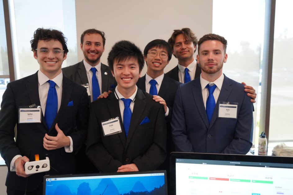 Six student stand together, all wearing black suits with a white shirt and blue tie.